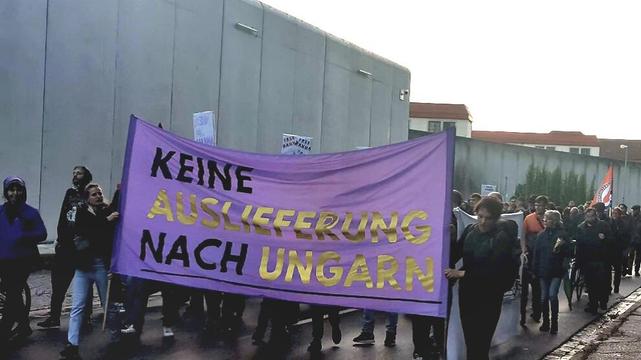 Foto des Protests vor der jva Nürnberg. Mehrere Menschen laufen hinter einem lila Banner auf welchem in schwarz Gold „Keine Auslieferung nach Ungarn“