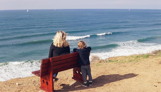 Unsplash photo from Tel Aviv, Israel showing the backs of a mom with blond hair and her young kindergarten-aged son on a sandy beach, gazing out over the sea. She is sitting on a red wooden bench placed just at the water's edge. He is standing behind, and leaning his arms over the top of the bench. In the distance are 3 tiny white sailboats. Mood is a joyful family snapshot on a nice day by the water, perhaps thinking about what there may be out beyond it. 
Captioned, "Overlooking great surf in TLV with Sophie and Tom". 

Tel Aviv Israel, tel-aviv, Israel 
Dated 2018 Dec 08, by Chen Mizrach. 
www.Unsplash.com/photos/woman-sitting-on-bench-near-boy-standing-at-the-back-7PcjhCWMwpM 

www.KronoMoon.org 🏳️‍🌈 ☮️ 
#KronoWatch