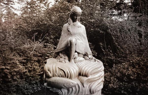 A stone sculpted statue of a Coast Salish Indigenous woman with long hair, draped in a shawl, kneels with one leg bent on a sculpted base resembling water, with salmon swimming. Ducks are at her feet. It is surrounded by lush greenery, giving a serene and natural scene.