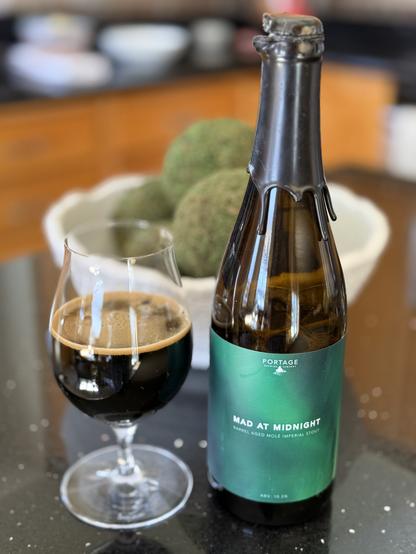 A snifter glass of dark beer. The tall brown bottle is to the right with a dark green label and white lettering. A bowl with green balls a counter fill of dishes are in the blurred background- lunch time!