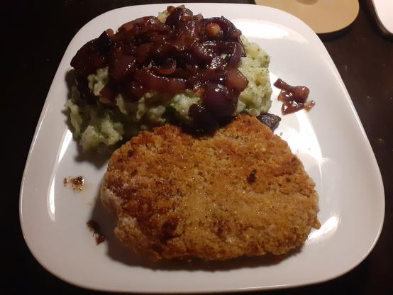 Paniertes Haferschnitzel liegt vorne auf einem weißen quadratischen Keramikteller, hinter dem Schnitzel liegt der Kartoffelstampf mit den karamellisierten Zwiebeln oben drauf.