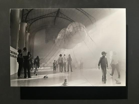 A black-and-white photograph of a balloon with a complex shape that floats inside a hangar, with people under it.