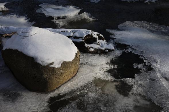A photo of a frozen brook. There is a large snow-covered rock on the left.