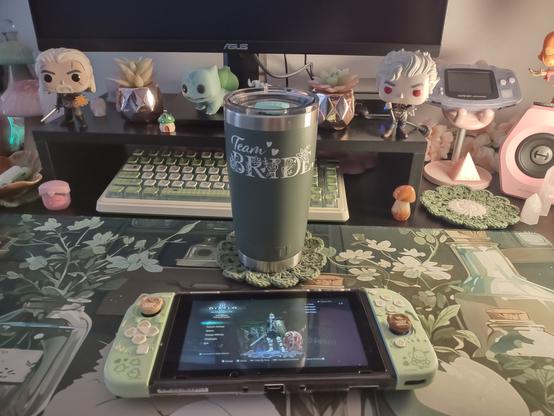 This is a photo of my gaming desk with sage and darker green shades, along with pops of pink. I have my Nintendo Switch sitting on my desk and resting behind it is my Yeti mug, also dark green. On it, it says Team Bride, which was a gift from one of the other bridesmaids from my best friend's wedding. I have Bulbasaur, Geralt, and Astarion Funko Pops beside my monitor, mushrooms everywhere, and crystals all around.