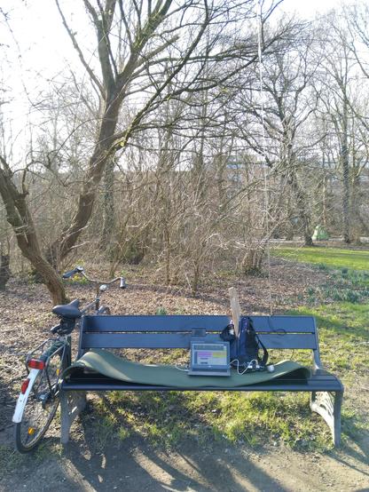 A bench in a park (Vondelpark in Amsterdam), with a backpack, laptop, a small QDX radio and an antenna tuner. Behind the bench is a vertical antenna. My bike leans against the bench.