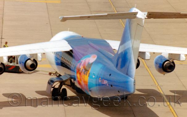 High rear view of a high-winged, 4 engined jet airliner being pushed back from left to right and towards the camera by a low-slung, dark blue truck on the left of the frame.
The front of the plane is white, while most of the fuselage isa deep blue, with the image of a planet on the rear fuselage, in a water-colour style.
Concrete apron fills the rest of the frame, with a yellow stripe running from the top to the bottom visible under the right wing.