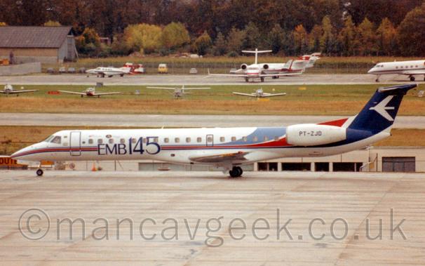 Side view of a long, thin, twin engined jet airliner with the engines mounted on the sides of the rear fuselage, taxiing from right to left.
The plane has a thin light blue, red, and dark blue stripe running along the body, getting wider as it sweeps up towards the rear fuselage and tail.
There are large blue "EMB145" titles on the forward fuselage, and the registration "PT-ZJD" in black on the engine pods.
Grey concrete apron fills the foreground, with a handful of light aircraft parked on grass in the background, leading up to another apron area filled with BizProps and BizJets, with trees beyond that.