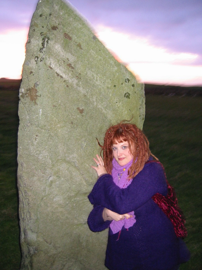 A young white woman with long red dreadlocks stands beside a large standing stone. The photo is slightly distorted in the gloaming, the distance appears to be spinning, the horizon blurs