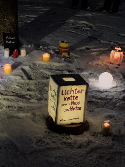 Einige Kerzen und Laternen, die im Schnee stehen. Im Vordergrund eine große Laterne, die mit "Lichterkette gegen Hass und Hetze" beschriftet ist. Im Hintergrund 3 Kerzen die vor einem schwarzen Schild stehen, dass mit "Amel" und "Hafsa" beschriftet ist.