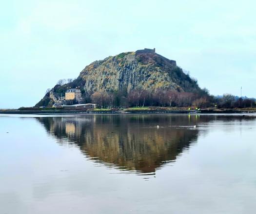 A large 330 million year old volcanic plug with a castle perched on top of of it reflected in the calm waters of the Clyde.