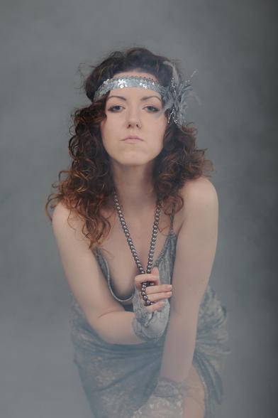 The image shows a person dressed in a vintage or Roaring Twenties-style outfit. They are wearing a sequined headband with a feather, a dress, and a long beaded necklace, along with lace gloves. The person's style is reminiscent of the 1920s fashion era.