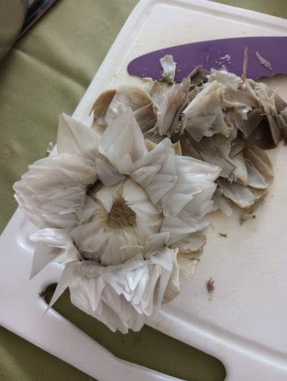 A steamed artichoke with its outer petals all removed sits on a cutting board with a knife, near some of the outer detritus. The very inner petals are folded out, so you can see a little bit of the inside of the flower, the "choke" part. It very much looks like a flower now, not a standard idea of a vegetable. Rather than be olive green like the outer petals, the inner petals are white-gray with a hint of purple. It's quite lovely.