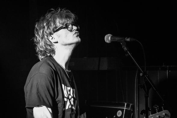 Landscape orientation. Black and white photo of a musician. He is blonde and has a mullet hairstyle. He is pursing his lips in front of a microphone. It is a head and shoulder shot