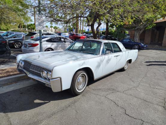 A pale blue or white 1962 Lincoln Continental 4-door is parked under a tree at Billy Reed's Restaurant in Palm Springs California. It is seen from the driver's side in  a front three quarter view. 

All the windows are open, revealing the light blue leather interior.