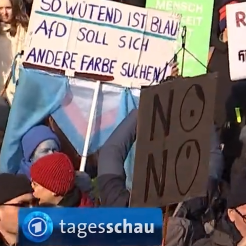 Ein Ausschnitt aus einem Screenshot der tagesschau (das Logo ist unten links in der Ecke zu sehen). Im Vordergrund rechts ein Demoschild aus Pappe, auf dem übereinander zweimal NO steht. 
Das O vom oberen NO zeigt einen stilisierten Hitler mit Seitenscheitel und Bärtchen, das untere O einen stilisierten Merz mit seiner typischen Glatze.
Im Hintergrund ein weiteres Schild mit der Aufschrift: So wütend ist blau: AfD soll sich eine andere Farbe suchen.