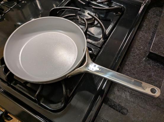 A small fry pan rests on a burner on a gas stove. The fry pan has a grey ceramic coating and a stainless-steel handle.