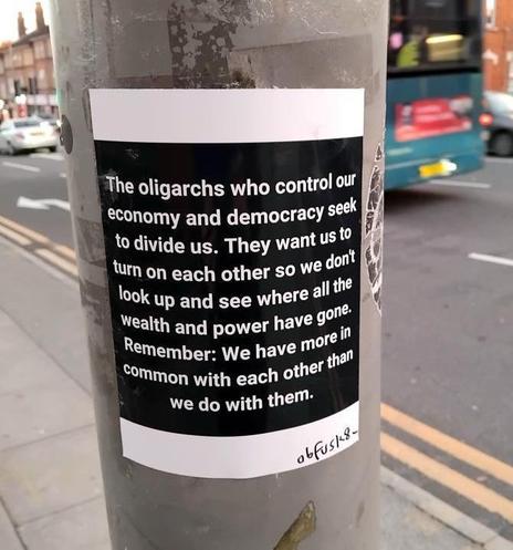 A black-and-white political sticker on a metal pole in an urban setting, featuring bold white text on a black background. The message criticizes oligarchs for controlling the economy and democracy, alleging that they seek to divide people to distract from wealth and power concentration. It ends with a unifying statement: "We have more in common with each other than we do with them." The sticker appears slightly peeling at the bottom, with a handwritten tag “obfusc-18” in the corner. A blurred city street with a bus and buildings is visible in the background.