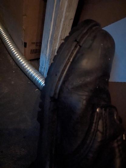 A close up of a cracked black boot on my right foot, next to a table leg, a loose spare piece of flexible metal conduit and a cardboard box on a mucky dark grey coloured floor.