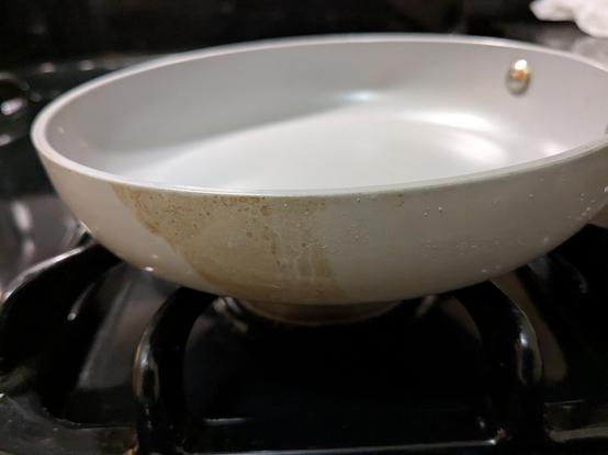 Brown discolouration is seen on the outer surface of a grey fry pan that is resting on a gas stove burner. The discolouration is where hot butter dripped down the outside of the fry pan.