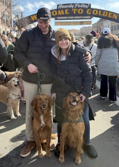 Man and woman and their golden retrievers , all facing the camera, beneath the Howdy Folks! Welcome to  Golden arched street  sign with other golden retrievers and companions in the street background.