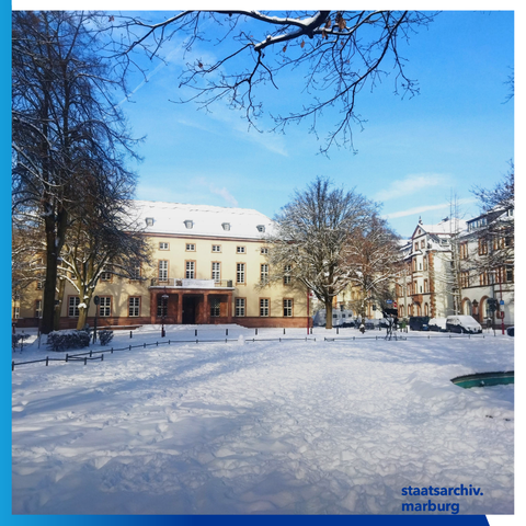 Schnee vor dem Haupteingang des Staatsarchivs in Marburg am Friedrichsplatz. Rechts im Bild ein Teil vom Brunnen. Vor dem Gebäude stehen links und rechts zwei Bäume.