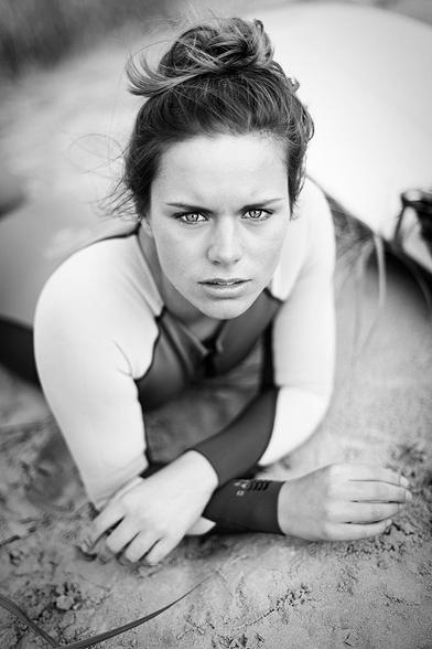 Surfer girl lying prone on a dune and looking directly into the camera with her upper body raised.