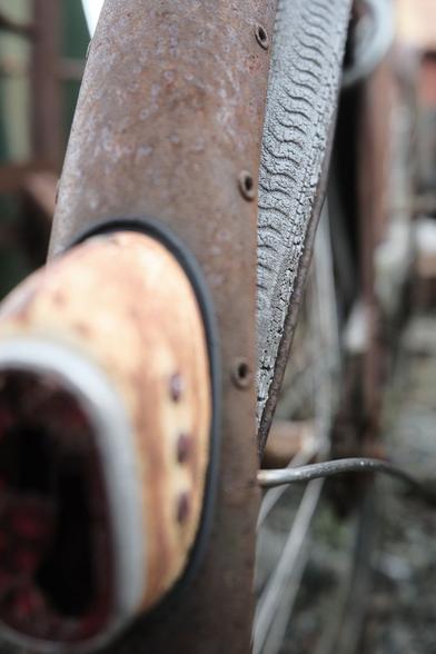 Närbild bakifrån på en rostig och sned bakskärm på en cykel med ett lika snett och torkat däck. Baklampa närmast i bild är ur fokus, precis som ekrarna och navet längst bort i bilden.