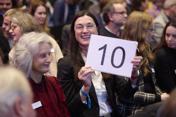 Rating bei einem Science Slam: Eine lachende Frau hält eine Stimmkarte mit einer 10 (Bestnote) hoch. Ringsum weitere Besucherinnen und Besucher im Gespräch miteinander.