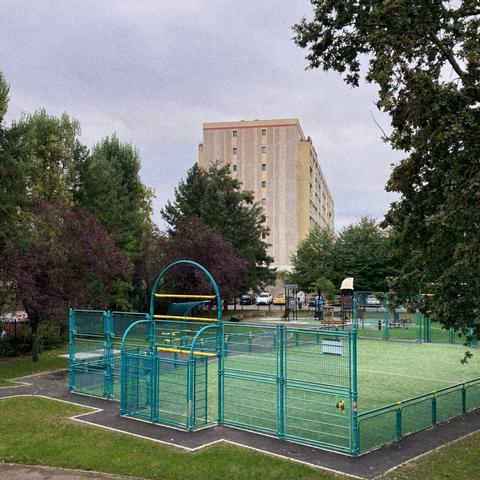 Un parc avec un terrain de foot et divers jeux vide devant à proximité d'un grand immeuble. Photographie en couleurs.