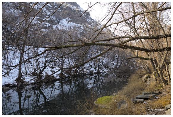 A slow river flowing diagonally through the frame. Both banks have trees which branches lean over the water. The left bank is illuminated by the sun, and has almost no snow, as opposed to the right bank where the sun doesn't shine enough to melt the snow.