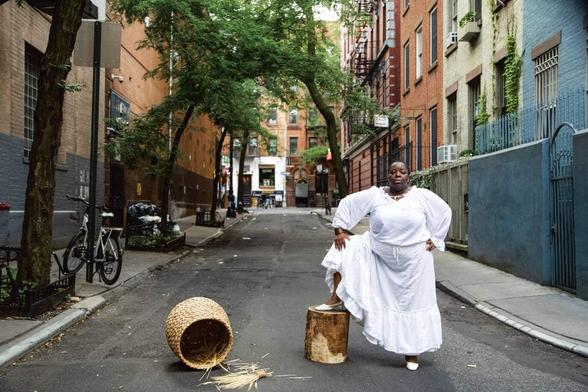 Nona Faustine se tien au milieux de la rue, un pied posé sur un objet cylindrique, en robe blanche et escarpins blancs.