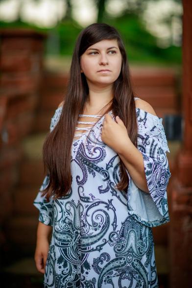 Eine junge Frau mit langen braunen Haaren steht in einem idyllischen Schlossgarten. Sie trägt ein romantisches weißes Sommerkleid mit blauem Paisley-Muster und blickt verträumt in die Umgebung.