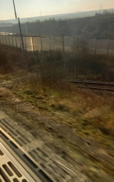 Slightly blurry view outside a train window onto a double fence with narbed wire and some greenery in sunny weather