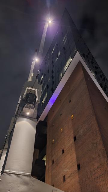 Aus der Froschperspektive bei Nacht. 
Die Ecke eines Backteingebäudes mit überspitztem Winkel, links neben dem Gebäude steht ein alter, weißer Hafenkran.
Im unteren Teil des Gebäudes,der aus rotem Backstein besteht, sind übereinander fünf Reihen je drei kleiner Fenster zu sehen, die mittlere ist beleuchtet. Der obere Teil des Gebäudes besteht aus Glas, auch hier sind einige Fenster beleuchtet. Der Hafenkran verfügt über drei nach unten gerichtete Leuchten. Sie sind im Abstand eines ungefähren Krandrittels übereinander angebracht.