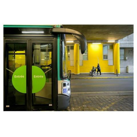 Un bus vert et noir est stationné à gauche de l’image, ses portes arborant deux grands cercles verts avec l’inscription « Entrée ». En arrière-plan, un mur jaune vif avec des colonnes et un passage sous un pont. Une personne pousse un fauteuil roulant tandis qu’une autre semble tracer un geste dans l’air, créant une scène dynamique et intrigante.