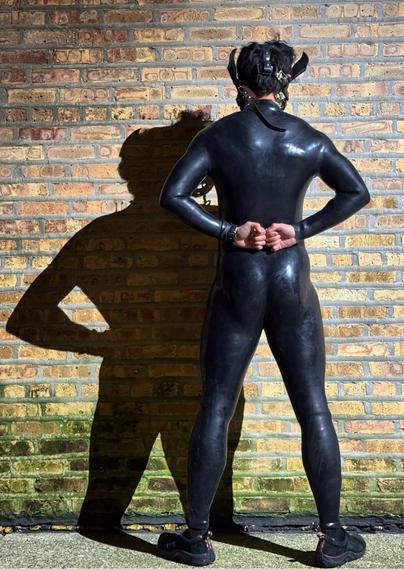 Under the glow of a stark light, a figure stands against a textured brick wall, their shadow stretching dramatically to the side. Encased in a sleek black silicone bodysuit, every curve and contour is accentuated. The material reflects the light subtly, giving it a polished, almost liquid-like sheen. Their stance is confident yet restrained, with hands clasped behind their back, exuding a sense of quiet power and introspection.

The addition of a leather hood with pointed equine ears adds an air of mystery and intrigue, blending elements of playfulness with bold self-expression. The hood's straps and buckles provide texture and contrast against the smooth neoprene, hinting at a narrative of individuality and freedom within boundaries.

The backdrop—a weathered brick wall—offers a gritty, urban aesthetic that juxtaposes with the futuristic and tactile appeal of the outfit. The muted tones of the bricks allow the black suit to command attention, while the shadow creates depth and dimension in the composition.