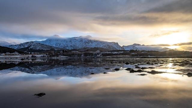 The image shows a scenic landscape with snow-covered mountains in the background. There is a body of water in the foreground reflecting the mountains and the sky. The sky appears to be overcast with some sunlight breaking through, creating a tranquil and picturesque scene.