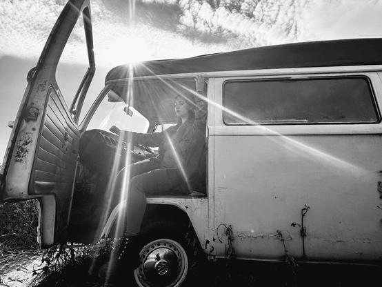 A black & white photo of a dark, long haired woman, sitting in the drivers seat of a vintage van with the door open. Sunlight creates a starburst effect through the window & textured sky.