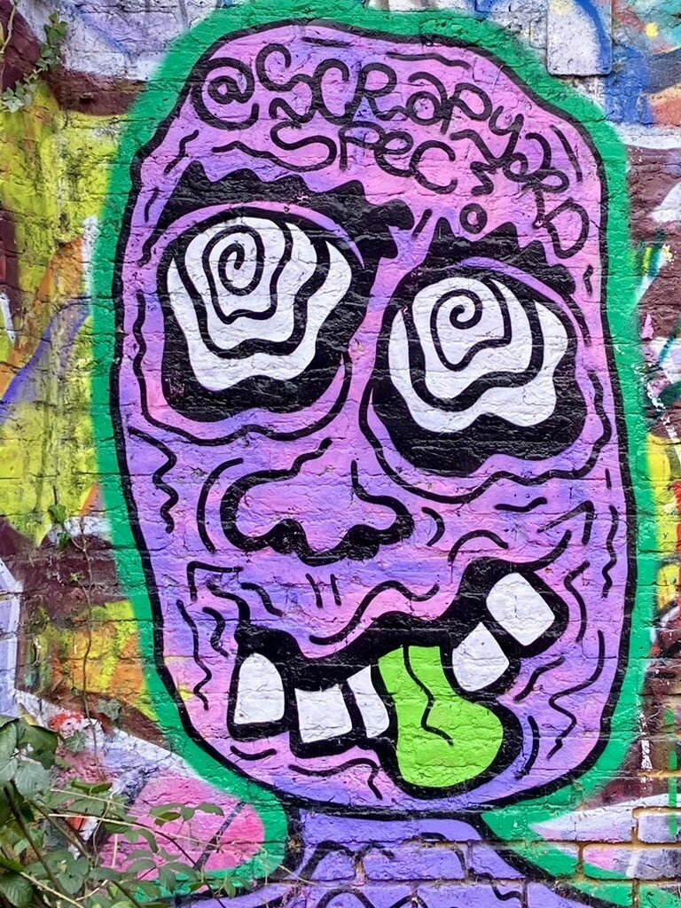 Street art on a brick wall that has other street art layers behind it: a swirly purple face with swirly white eyes and a green tongue sticking out with the words "@scrapyard spec" written on its forehead.