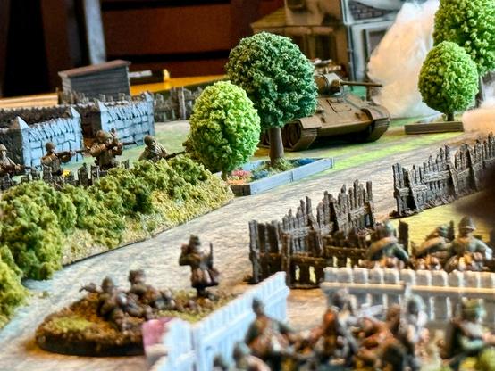 Photograph of a tabletop war game set World War II. A large enemy tank is rounding a tree while friendly infantry huddles behind wooden fences in the foreground.