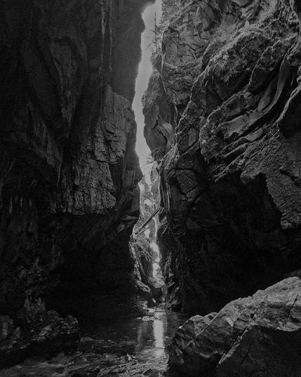 A narrow canyon with steep, jagged rock walls towering above a shallow creek. Sunlight illuminates a narrow passage between the rocks, creating a stark contrast with the darkness of the canyon.