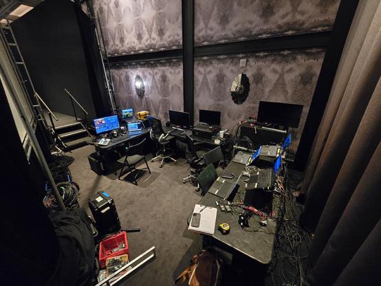 Backstage area with multiple computer workstations, monitors, cables, and equipment on tables. A staircase is visible on the left, and a notebook and measuring tape are on one table.