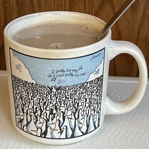 Photo of a Mug of tea with a far side cartoon on it: a tight huddle of penguins that stretches as far as the eye can see against an arctic landscape. One penguin is stretched up above the rest and singing “I gotta be me, oh, I just gotta be me… “