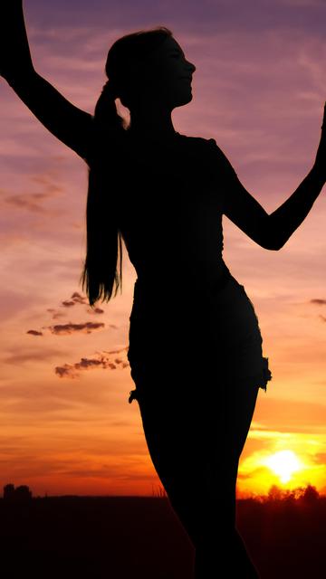 The image shows the silhouette of a person standing against a colorful sunset or sunrise sky. The person appears to have their arms extended and is standing in a natural landscape.