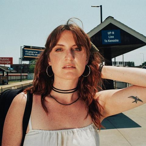 A white woman with auburn hair stairs directly into the camera. She has bangs along her forehead and is wearing hoop earrings. She has a black backpack hung over her shoulder and is wearing a black choker necklace and white tank top. She is standing on the platform of an outdoor railroad station.