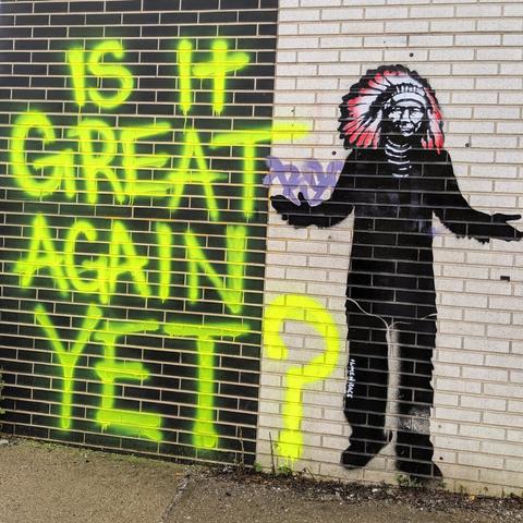 Yellow graffiti text on a brick wall reads "is it great again yet?", as an elder Native American in a traditional headdress shrugs with extended arms.