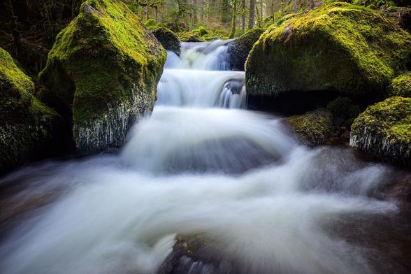 Zu sehen ist eine Wasserfallszenerie mit einer großen Kaskade im Mittelpunkt, während sich an den beiden Seiten riesige, bemooste Steine befinden, die mit Eiszapfen behangen sind.