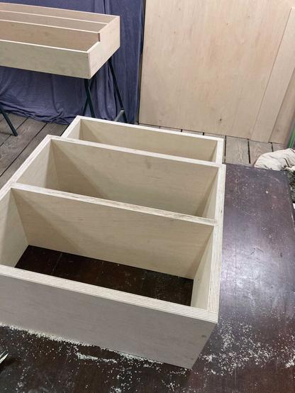 a picture of my studio. in the front a birch plywood shelf is on a dark brown table.
in the back another birch plywood shelf is lying on some black table trestles in front of a blue fabric.