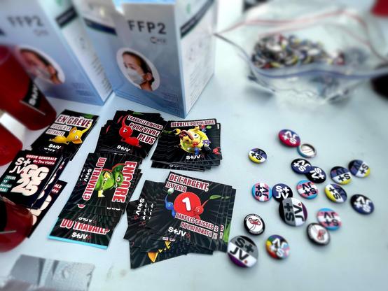 Photo du rassemblement du STJV à Montpellier, dans le cadre de la grève générale du jeu vidéo du 13 février 2025. Une table est couverte de goodies STJV. À gauche, des gobelets rouges avec un dessin de drapeau STJV noir. En haut des boîtes de masques FFP2. En haut à droite un sachet plastique contenant des badges STJV. En bas à droite des badges STJV trans, LGBT, assexuel, anarcha-féministe, non-binaire et antifascist game worker. Au milieu des stickers STJV Pikmin "en grève jusqu'à la retraite", "travailler moins pour vivre plus", "révolte populaire", "de l'argent il y en a des les caisses du patronat", des stickers STJV Warcraft 3 "Encore du travail ?", et des stickers STJV GG25 Grève général du jeu vidéo.