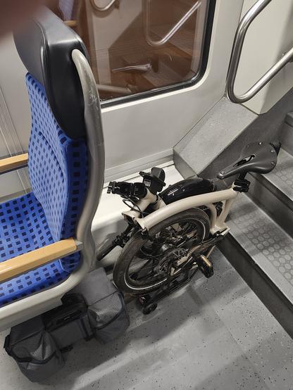 A folded Brompton G Line bicycle between a seat and the stairs in a commuter train.